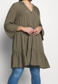 Khaki tunika s výstřihem do V, tříčtvrtečním zvonovým rukávem a vícestupňovým volánkovým lemem. Vyrobena z měkké látky s hladkým povrchem.