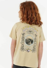 Camiseta gráfica beige con una imagen central redonda enmarcada por calaveras ilustradas y figuras parecidas a tótems. Texto "Ripcurl" en letras coloridas arriba.