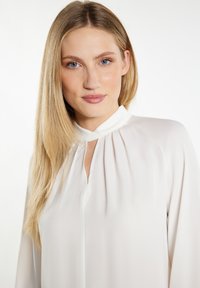 Weiße Bluse mit steilem Kragen und pleite Front, ausgestattet mit einer Schlüsselkerbe. Hergestellt aus einem glatten, leichten Stoff mit weicher Textur.