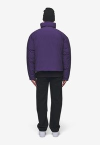 Fioletowa puchowa kurtka z podniesionym kołnierzem, nosząca czarne spodnie i czapkę beanie. Charakteryzuje się gładką fakturą i skróconym krojem. Białe sneakersy.