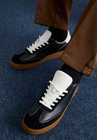 Black and white low-top sneakers made of leather and suede, featuring white laces, a rubber gum sole, and a logo on the tongue.