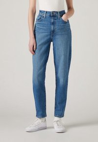 Personne portant un jean bleu taille haute à coupe droite, un haut blanc, des chaussettes blanches et des baskets blanches à lacets, debout sur un fond clair.