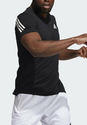 Mann in schwarzem Adidas-Shirt mit drei weißen Streifen am Ärmel und weißen Shorts in Bewegung vor einfarbigem Hintergrund.