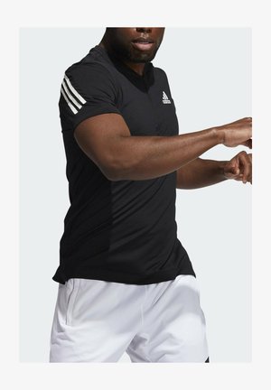 Homme en t-shirt noir Adidas avec trois bandes blanches sur la manche et short blanc en mouvement sur fond uni.