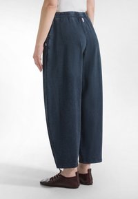 Osoba nosząca luźne ciemnoniebieskie spodnie z elastycznym pasem i ciemnoczerwone buty na sznurowadła, stojąca na białym tle.