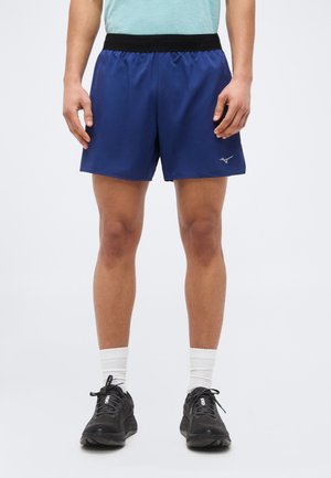 Άνδρας που φοράει ναυτικό μπλε αθλητικό σορτς με μαύρη μέση, λευκές κάλτσες και μαύρα αθλητικά παπούτσια, στέκεται μπροστά σε ανοιχτόχρωμο φόντο.