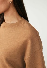 Ruskea huppari, jossa on pyöreä kaula-aukko, ribbaustyölö ja pehmeä, teksturoitu kangas. Ominaisuuksiin kuuluvat roikkuvat hartian saumat ja rento istuvuus.