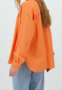 Vrouw met golvend haar draagt een losse, oranje top met lange mouwen en lichtblauwe spijkerbroek, gezien van achteren tegen een effen lichte achtergrond.