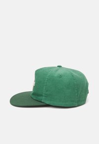 Gorra de béisbol de pana verde con visera plana, que presenta un botón de tela en la parte superior y un logo bordado en blanco en el frente.