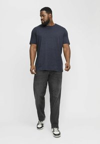 Marineblå t-shirt med korte ærmer, løs pasform, parret med mørkegrå jeans og sort-hvide sneakers, stående mod en ensfarvet baggrund.
