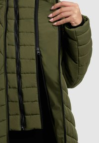 Giacca imbottita verde oliva con un motivo trapuntato, che presenta molteplici cerniere nere e un colletto regolabile. Design del colletto alto per garantire calore.