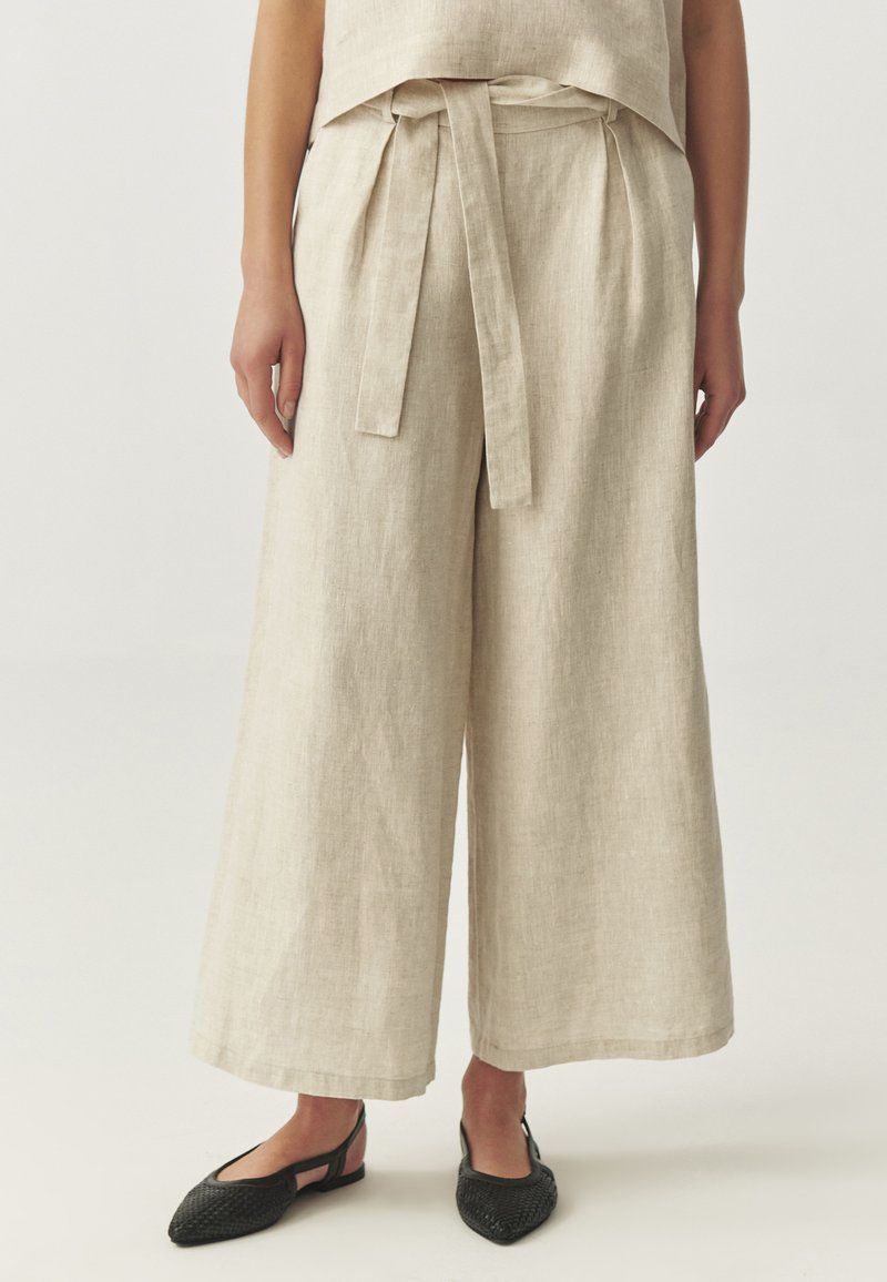 Pantalons larges en lin de couleur beige clair, dotés d'une taille ceinturée, de poches latérales et d'un design fluide. Assortis avec des chaussures slingback noires.