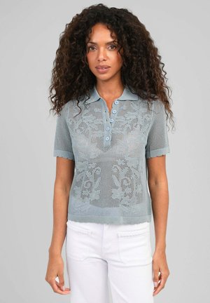 Femme aux longs cheveux bouclés porte un polo en maille bleu clair à manches courtes avec motif floral et un pantalon blanc sur fond gris.