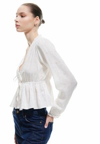 Hvid bluse med lange ærmer og struktureret stof, med en knytterne lukning, indsnævret talje og blomstret hulmønster. Sammen med mørke jeans.