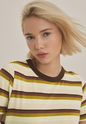 Blondýna s krátkými vlasy, která má na sobě proužkované tričko v odstínech hnědé, žluté a krémové, a dívá se mírně dolů.