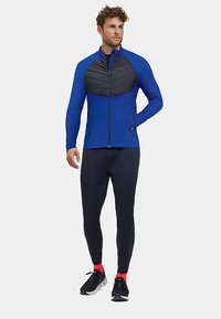 Blue athletic jacket with black padded chest panel, zip closure, two side pockets. Worn with black leggings and red-accented shoes.