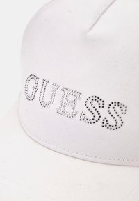 Cappello da baseball bianco in cotone, con logo "GUESS" arricchito da strass trasparenti, forma strutturata e dettagli cuciti.