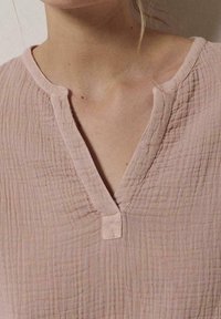 Blusa rosa chiaro con tessuto strutturato, scollatura a V e un piccolo bottone frontale. Materiale morbido e stropicciato con un motivo sottile.