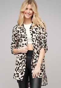 Beige Hemd mit Knöpfen und schwarzem Leopardenmuster, mit hochgeklappten langen Ärmeln, über einem weißen Cropped-Top und schwarzen hochgeschnittenen Hosen getragen.