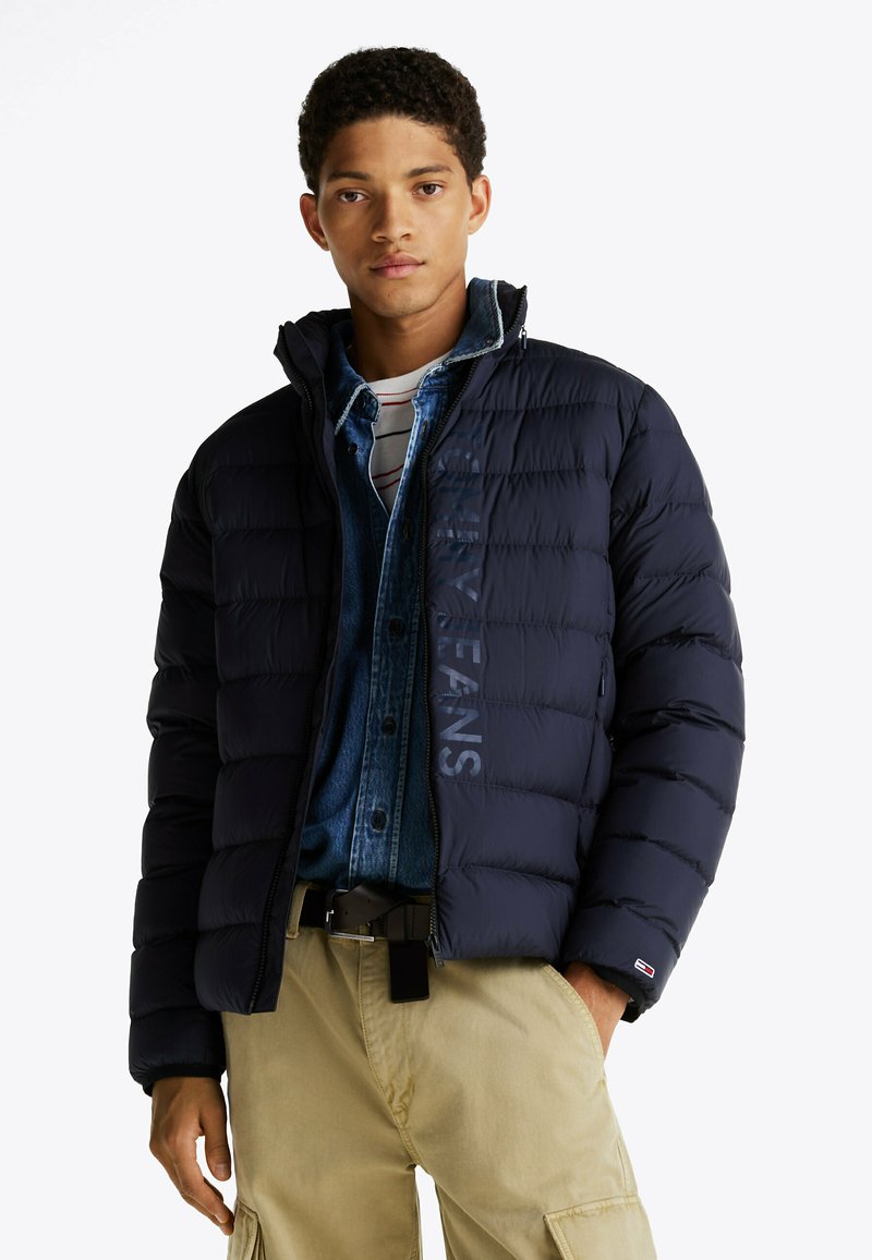 Marine blauwe pufferjack met een ritssluiting, voorzien van gestikte panelen en logodetail. Geraadpleegd over een denim shirt, gecombineerd met beige cargobroek.