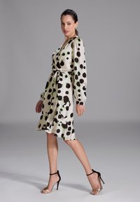 Robe portefeuille en satin avec un fond blanc et de grands pois noirs et verts. Elle dispose de manches longues, d'une ceinture à nouer et d'un ourlet volanté.