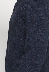 Gros plan sur une personne portant un pull en maille bleu marine texturé, montrant l'épaule, le haut du bras et la poitrine.