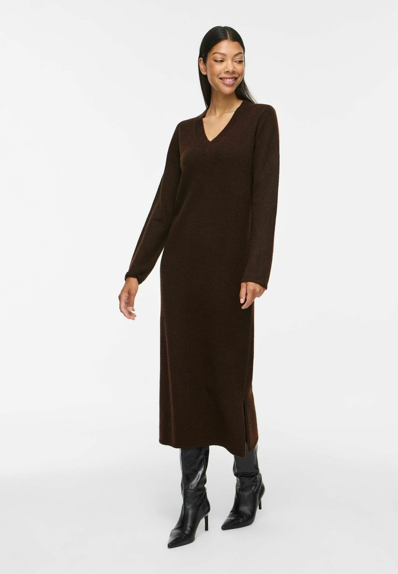 Femme souriante portant une longue robe en maille marron foncé à manches longues et des bottines noires à talons sur un fond blanc uni.
