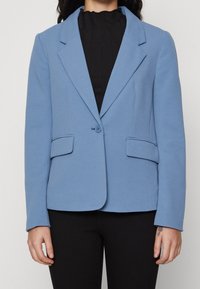 Světle modrý blazer vyrobený z texturované látky. Obsahuje klasický klopový límec, dvě přední kapsy a zapínání na jeden knoflík. Kombinovaný s černým topem.