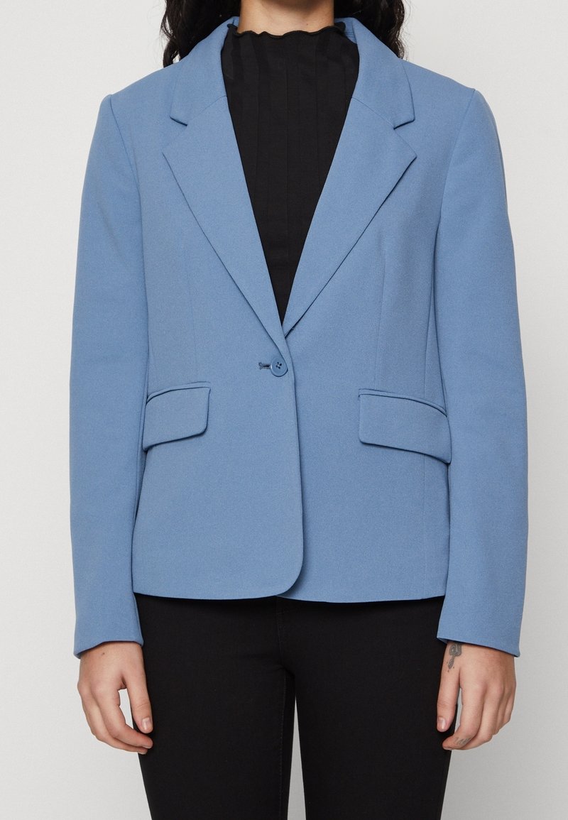 Světle modrý blazer vyrobený z texturované látky. Obsahuje klasický klopový límec, dvě přední kapsy a zapínání na jeden knoflík. Kombinovaný s černým topem.