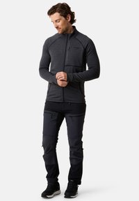 Herren-Midlayer-Jacke in Dunkelgrau mit Reißverschluss, strukturiertem Stoff und zwei Seitentaschen, kombiniert mit schwarzen Outdoor-Hosen.
