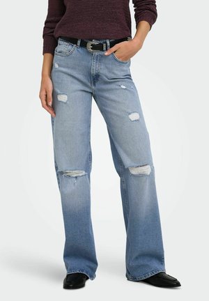 Personne portant un jean large déchiré bleu clair, une ceinture noire, un pull violet foncé et des chaussures noires, debout avec une main dans la poche.