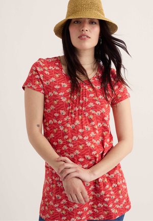 Femeie purtând un top roșu cu flori și mâneci scurte și o pălărie tip găleată țesută, culoare bej, având părul închis la culoare și mâinile încrucișate în față.