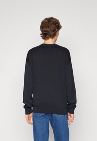 Schwarzer Pullover mit Rundhalsausschnitt, langen Ärmeln und gerippten Bündchen. Die Rückseite verfügt über eine glatte Textur, kombiniert mit blauen Jeans.