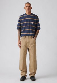 Μπλε ναυτικό ριγέ μπλουζάκι με μικρό λογότυπο, συνδυασμένο με μπεζ παντελόνια εργασίας με εμφανείς ραφές. Μαύρα παπούτσια ολοκληρώνουν την εμφάνιση.