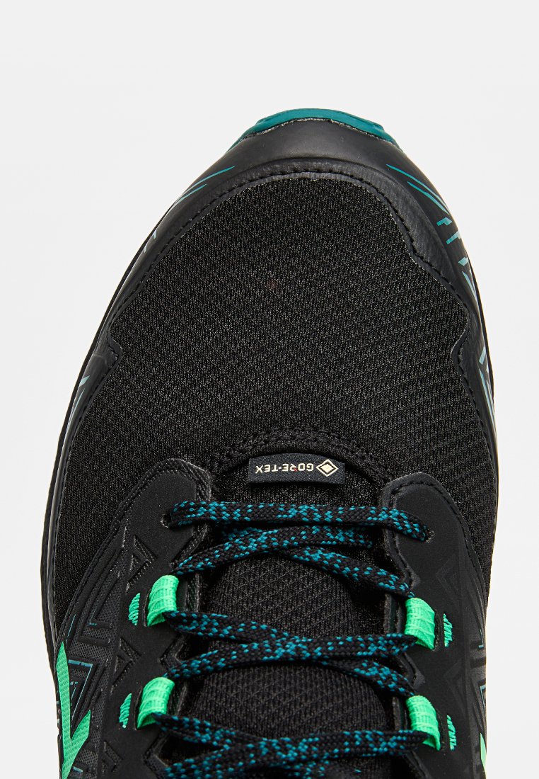 Zapatilla deportiva negra con un upper de tela texturizada, acentuada por detalles verdes y cordones con un diseño a rayas. Cuenta con un logo prominente y un diseño de caña alta.