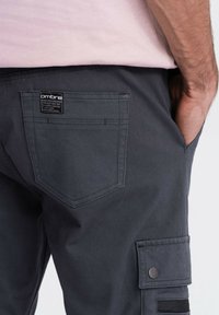 Muž oblečený v tmavě šedých cargo kalhotách s bočními a zadními kapsami, jedna ruka částečně v boční kapse a světle růžové tričko zasunuté dovnitř.