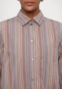 Personne portant une chemise à col et manches longues avec des rayures verticales multicolores et des boutons dépareillés, dont des boutons dorés, noirs et gris.