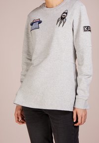 Szara bluza wykonana z miękkiego materiału, z naszywkami przedstawiającymi rakietę i kapelusz, z okrągłym dekoltem i długimi rękawami.