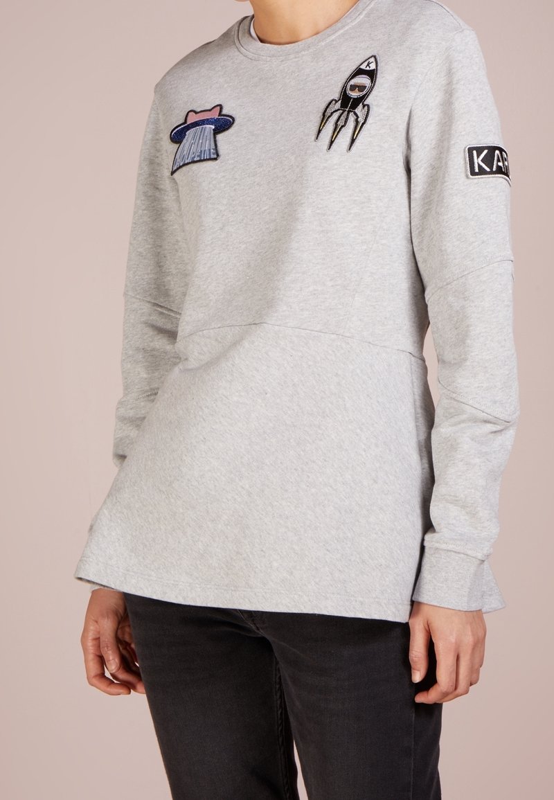 Szara bluza wykonana z miękkiego materiału, z naszywkami przedstawiającymi rakietę i kapelusz, z okrągłym dekoltem i długimi rękawami.