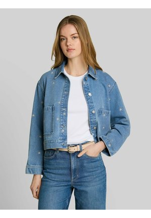 Junge Frau, die eine hellblaue bestickte Jeansjacke über einem weißen Hemd und blauen Jeans trägt, steht mit einer Hand in der Tasche.