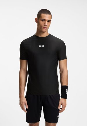 Junger Mann, der ein schwarzes Sportshirt, Shorts und ein Armband trägt, alle mit dem "BOSS"-Logo, steht vor einem einfarbigen weißen Hintergrund.