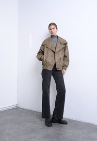 Γυναίκα στέκεται ανάμεσα σε λευκό τοίχο, φορώντας υπερμεγέθη καφέ σακάκι, γκρι ζιβάγκο, μαύρο τζιν με φαρδιά πόδια και μαύρα μοκασίνια, με τα χέρια στις τσέπες.