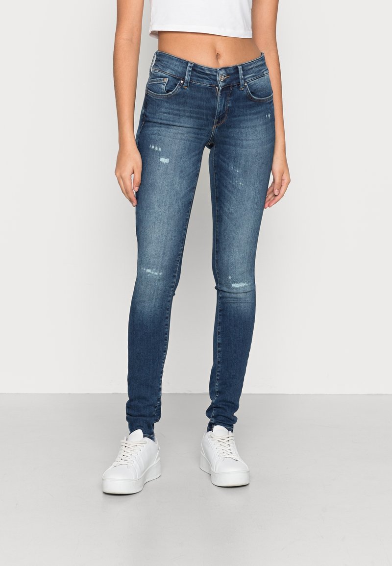 Skinny-fit spijkerbroeken in donkerblauw met lichte distressed details, vijfzakkenontwerp en ritssluiting, gecombineerd met witte sneakers.