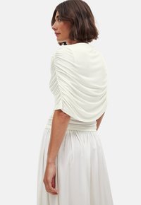 White top featuring gathered fabric along the sleeves and back, with a fitted waist and flowing skirt, made from lightweight, smooth material.