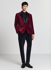 Blazer in velluto bordeaux con rever in raso nero, vestibilità sartoriale, due tasche laterali, abbinato a pantaloni neri e scarpe eleganti nere lucide.