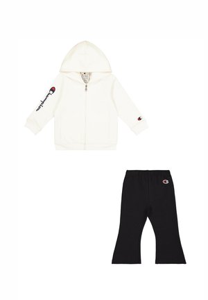 Sudadera blanca con cremallera y capucha, junto con pantalones negros, ambos con acentos del logo de Champion. Tejido suave, corte holgado con perneras acampanadas.
