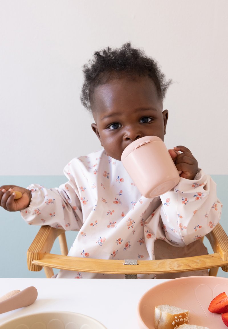 Un bavaglino lungo con un motivo floreale morbido indossato da un bambino piccolo, che tiene in mano una tazza rosa. È visibile un vassoio con posate rosa e alimenti.