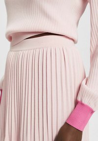 Haut en tricot côtelé rose pâle et jupe plissée, avec des poignets côtelés roses contrastants. Texture douce, taille ajustée et plis verticaux prononcés.