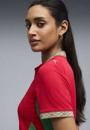 Mujer de cabello largo y oscuro, vestida con una camiseta polo deportiva roja y verde con cuello y ribetes de las mangas estampados, mirando por encima del hombro.