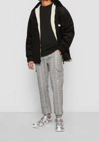 Mężczyzna w czarnej polarowej kurtce z sztruksu, czarnej koszuli, szarych spodni cargo w kratę i srebrnych sneakersach stoi na tle w odcieniu szarości.